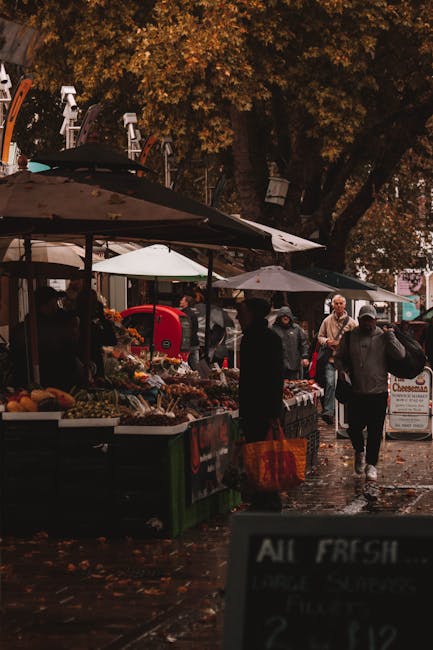 A bustling outdoor market scene in a small garden or park area, featuring multiple vendor stalls shaded by large umbrellas, with fresh produce and flowers displayed on tables. The ground appears damp,