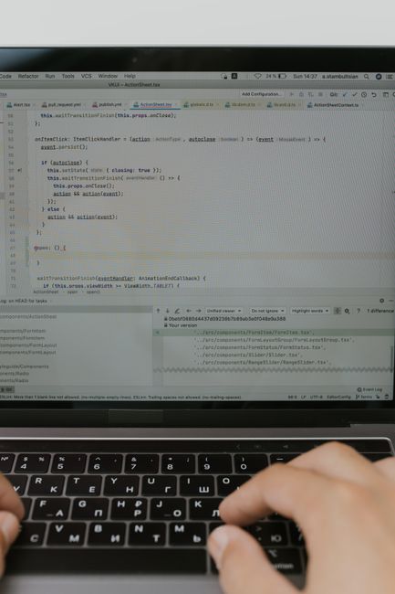 A close-up view of a person’s hands typing on a laptop keyboard in a well-lit outdoor garden setting. Behind the hands, the laptop screen displays lines of code, indicating ongoing digital work. In